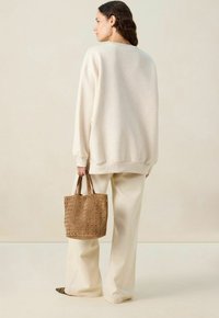 Sweatshirt beige oversize avec des poignets côtelés, associé à un pantalon large crème. Tenant un sac en toile perforée tan avec des découpes rondes.