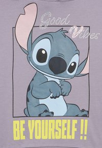 Tricou lavandă cu un personaj de desen animat cu urechi și ochi mari, cu text imprimat "Good Vibes" și "BE YOURSELF !!" în culoarea galbenă.