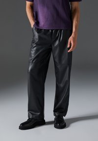Pantalones de cuero negro con un corte holgado, que presentan paneles cosidos y una textura suave. Combinados con zapatos negros y una blusa morada.