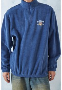 Marinblå fleece-pullover med hög krage, fjärdedels dragkedja och elastiska manschetter. Har en vit broderad logotyp på bröstet.