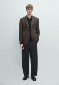 Blazer marrone su misura sopra una polo nera, abbinato a pantaloni neri larghi. Tessuto liscio, design strutturato e accenti minimalisti.