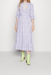 Žena v levandulových květinových midi šatech s volánovým výstřihem a průsvitným krajkovým vsazením, doplněných limetkově zelenými botami na jehlovém podpatku.