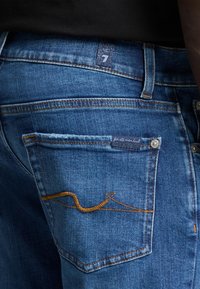 Jeans in denim blu con una tasca posteriore con cuciture arancioni e un'etichetta. Il materiale è denim classico con una texture liscia.
