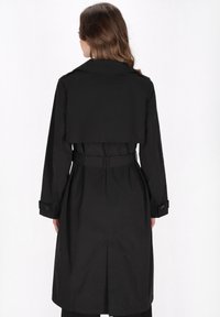 Fekete trench kabát övvel a deréknál, hosszú ujjakkal és hajtott gallérral. Közepes hosszúságú, és hátsó szellőzővel rendelkezik a szabad mozgás érdekében.