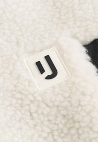 Hvid fleece-stof med en blød, tekstureret overflade. Har et rektangulært logo-patch med et sort "J" på en hvid baggrund.