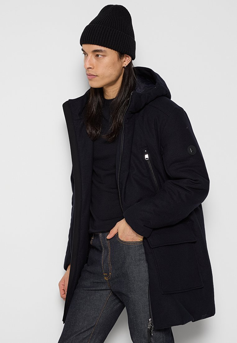 Dunkelblauer Parka mit Reißverschluss vorne, Kapuze und mehreren Taschen. Hergestellt aus strukturiertem Stoff. Kombiniert mit einem schwarzen Pullover und dunklen Jeans.