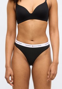 Črni komplet modrčka in spodnjic s gladko teksturo. Spodnjice imajo bel elastični pas z natisnjenim logotipom blagovne znamke.