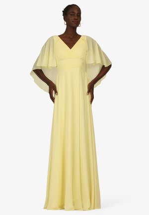 Frau in einem fließenden hellgelben Kleid mit plissiertem Oberteil und weiten Umhangärmeln, die mit Händen in der Hüfte vor einem weißen Hintergrund steht.
