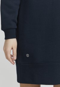 Mörkblå sweatshirtklänning med ribbade ärmslut, rak fåll och en liten silverlogotyp som accent vid nederkanten. Mjuk tygtextur.