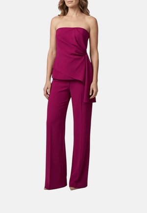 Donna che indossa un top magenta senza spalline con drappeggio laterale e pantaloni ampi abbinati, in piedi su uno sfondo semplice.