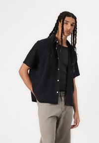 Black short-sleeve shirt in a relaxed fit, featuring a button-up design and soft denim texture. Paired with light gray pants.
