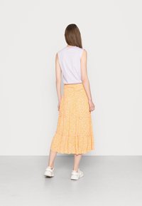 Nainen seisoo selkä kameraan päin, päällään hihaton vaaleanlila paita, oranssikuvioinen midi-hame ja valkoiset lenkkarit yksinkertaista taustaa vasten.