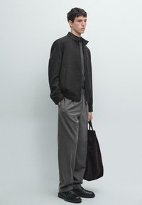 Ciemnoszara kurtka bomber, zielona koszula, jasnoszare spodnie, czarne buty oraz ciemna torba na ramię. Gładkie tekstury materiałów i minimalistyczny design.