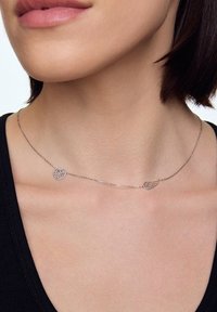 Srebrny naszyjnik z wiszącym wisiorkiem w kształcie serca i skrzydła, połączony delikatnym łańcuszkiem. Wisiorki prezentują skomplikowane wzory wycięte w metalu.