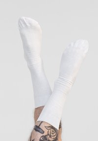 Weiße gerippte Socken mit gestrickter Textur, die ein klassisches Design aufweisen. Sichtbare Konturen der Beine und tätowierte Haut darunter.