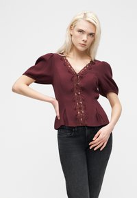 Νεαρή γυναίκα με ξανθά μαλλιά, που φοράει μπορντό μπλούζα με δαντέλα και μαύρα τζιν, ποζάρει με το ένα χέρι στη μέση πάνω σε λευκό φόντο.