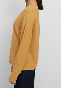 Maglione color senape con una texture a coste, maniche lunghe e un design con spalle leggermente scese. Abbinato a jeans scuri.