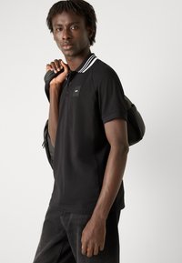 Camisa polo negra con tapeta de tres botones, cuello a rayas blancas y pequeño logo negro en el pecho; llevada con jeans oscuros y cargando una bolsa.