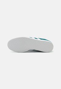 Shoe sole with a textured white rubber surface and a turquoise upper featuring three white stripes along the side. Flat, rounded shape.