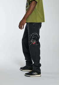 Los jeans de mezclilla negros presentan un destacado diseño bordado de un pantera en la pierna izquierda, combinados con zapatillas negras y una camiseta verde.