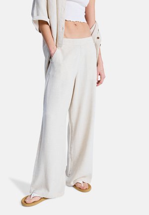 Helle beige Leinenhose mit weitem Bein, seitlichen Taschen, glatter Textur und lockerem Schnitt. Kombiniert mit einem weißen Crop-Top und Flip-Flops.