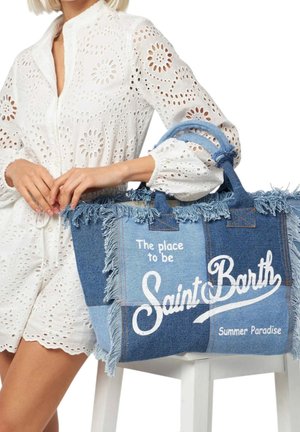 Femme en robe blanche à œillets tenant un grand sac cabas en jean bleu avec des franges et le texte « Saint Barth Summer Paradise ».