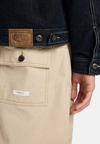 Giacca di denim con una toppa marrone e bottoni metallici, abbinata a pantaloni beige con tasca frontale e etichetta. Design casual e funzionale.