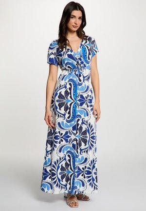 Donna con indosso un abito lungo bianco con motivi floreali astratti blu e blu scuro, maniche corte, scollo a V e sandali argentati con cinturini, in piedi.