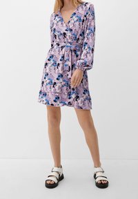 Robe florale violette à manches longues, décolleté en V profond, taille cintrée et ourlet à volants. Présente des accents bleus et noirs. Portée avec des sandales blanches.