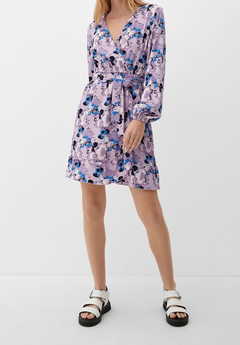 Robe florale violette à manches longues, décolleté en V profond, taille cintrée et ourlet à volants. Présente des accents bleus et noirs. Portée avec des sandales blanches.