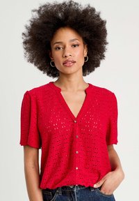 Blouse rouge à manches courtes avec un décolleté en V, présentant un motif texturé et des boutons sur le devant. Fabriquée en tissu léger et respirant.