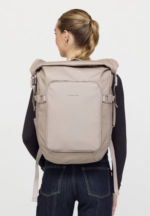 Vrouw met gevlochten haar draagt een beige rugzak met oprolbare bovenkant, een voorvak en verstelbare bandjes, gecombineerd met een zwarte top met lange mouwen en jeans.