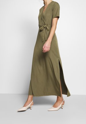Kobieta w oliwkowej sukience kopertowej z krótkimi rękawami, rozcięciem po boku i przewiązanym w talii, w połączeniu z białymi butami na niskim obcasie typu slingback.