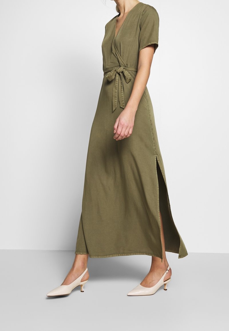 Femme portant une robe portefeuille vert olive à manches courtes, fente latérale et taille nouée, associée à des chaussures slingback blanches à petit talon.