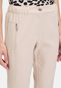 Beżowe spodnie sportowe z elastycznym pasem, przednimi kieszeniami na zamek i gładką teksturą. Charakteryzują się delikatnym połyskiem i dopasowanym krojem.