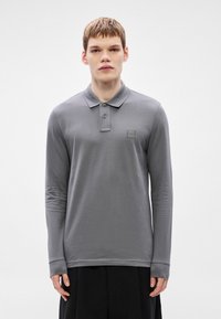 Graues Langarm-Poloshirt mit weicher Textur, zweiknöpfiger Knopfleiste und kleinem Markenpatch auf der Brust. Glatter Stoff, entspannte Passform.