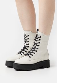 Botas de cuero blancas con cordones negros, suela de goma negra gruesa y detalles de costura. Altura hasta el tobillo con una pestaña en la parte posterior.