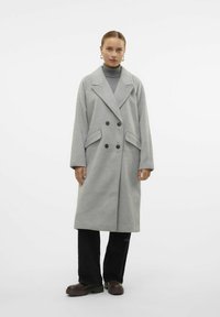 Cappotto grigio in lana doppiopetto con grandi bottoni, rever a L e tasche laterali. Abbinato con un dolcevita grigio e pantaloni neri.