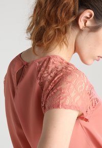 Brzoskwiniowa bluzka z koronkowymi rękawami, z zapięciem na guziki i detalu w postaci otworu na plecach. Gładki materiał o lekkiej, zwiewnej strukturze.