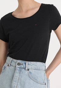 Frau trägt ein enges schwarzes T-Shirt mit kurzen Ärmeln, das in hochgeschnittene hellblaue Jeans gesteckt ist.