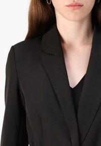 Svart blazer med en jevn tekstur, skarpe notched lapels og synlige sømdetaljer. Bært over en svart v-hals topp.