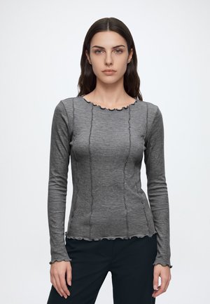 Giovane donna con capelli lunghi e scuri che indossa una maglia grigia aderente a manica lunga con dettagli di cuciture ondulate e pantaloni neri, in piedi davanti a uno sfondo bianco.