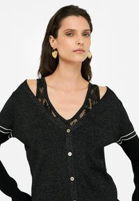 Cardigan nero e luccicante con scollatura a V profonda, chiusura con bottoni dorati e un top in pizzo sottostante. Presenta maniche lunghe con dettagli bianchi.