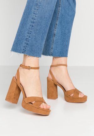 Pies con sandalias de tacón de bloque con plataforma de ante color beige y tiras en el tobillo, combinadas con vaqueros azules recortados.