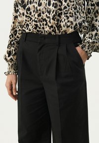 Black pleated trousers made of smooth fabric, featuring side pockets, paired with a patterned blouse with ruffled cuffs and a relaxed fit.