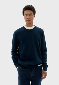 Pull ras du cou bleu marine avec un col et des poignets côtelés, superposé sur une chemise blanche, associé à un jean bleu. Tissu doux avec une texture lisse.