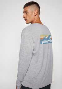 Svijetlosiva majica dugih rukava opuštenog kroja. Sadrži šareni dizajn oceanske valove i "patagonia" logo na leđima.