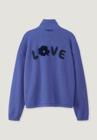Paarse fleece pullover met een hoge kraag, gekenmerkt door het woord "LOVE" in zwarte pluche letters en een zwarte bloemaccent op de achterkant.