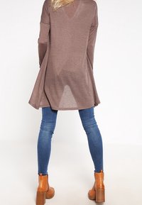 Dlouhá hnědá tunika s výstřihem do "V" a bočními rozparky, zkombinovaná s modrými úzkými džínami a hnědými kotníkovými botami na blokovém podpatku.