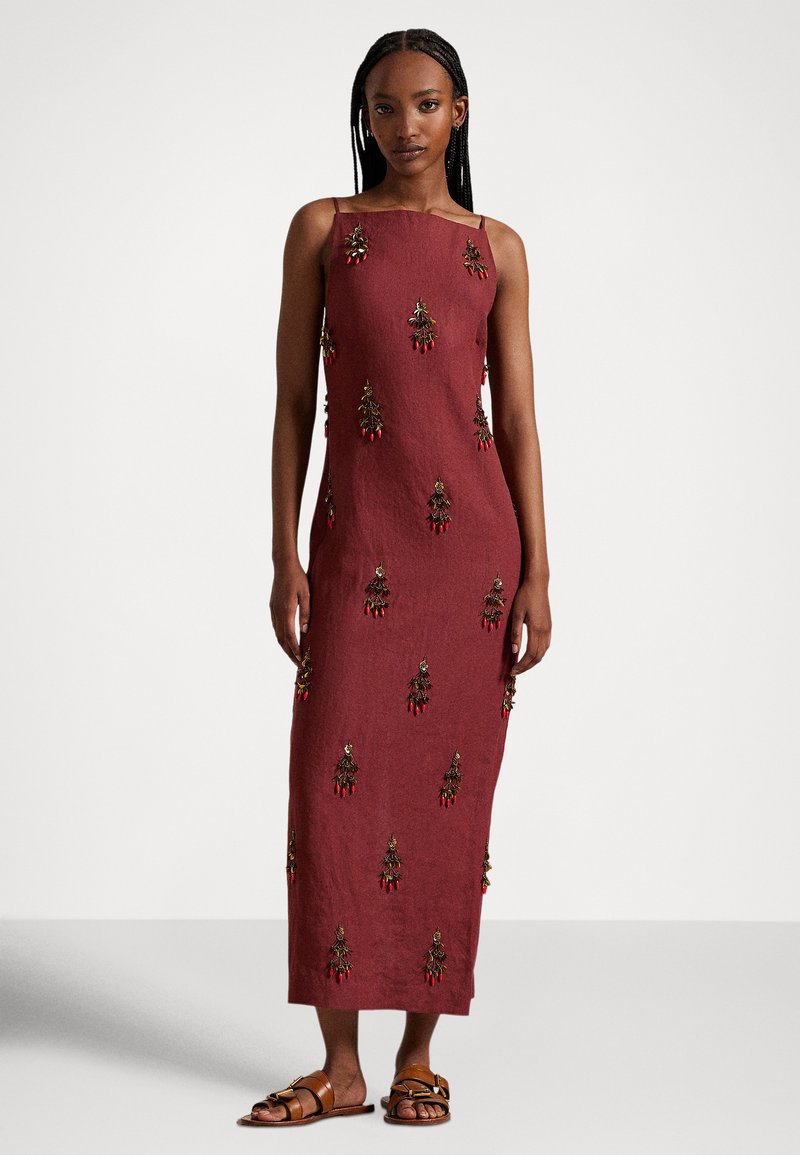 Frau in einem ärmellosen, bodenlangen, burgunderfarbenen Kleid mit Blumenstickerei und braunen flachen Sandalen, die vor einem schlichten hellen Hintergrund steht.